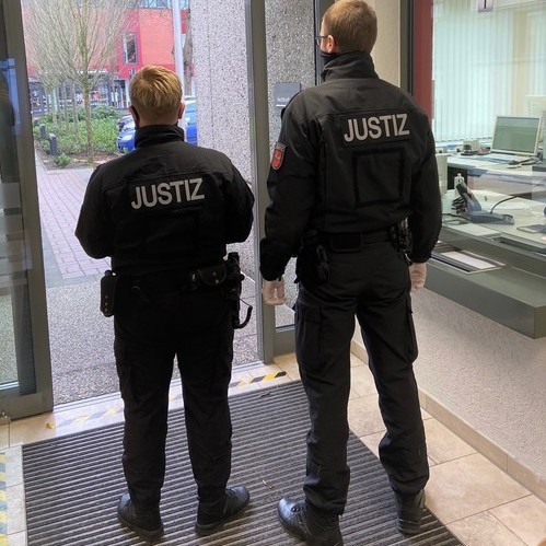 Eine Frau und ein Mann von hinten fotografiert. Jacken tragen Aufschrift Justiz.